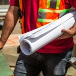 Construction workers in safety vests holding blueprints at a site during the day.